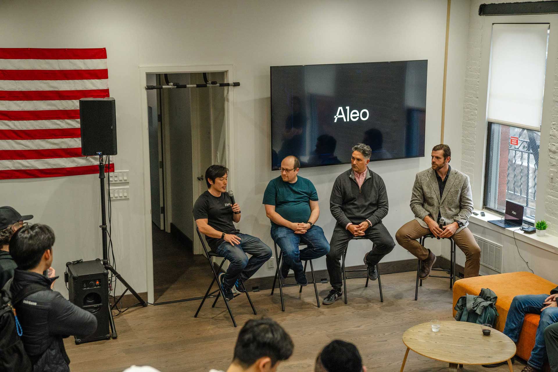 Four men sitting on stools in a panel discussion with Aleo logo on screen behind them.