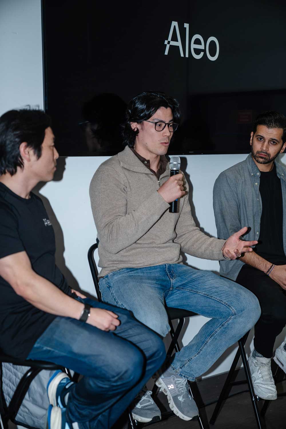 Three men seated in discussion, one speaking into a microphone with Aleo displayed on the screen behind.