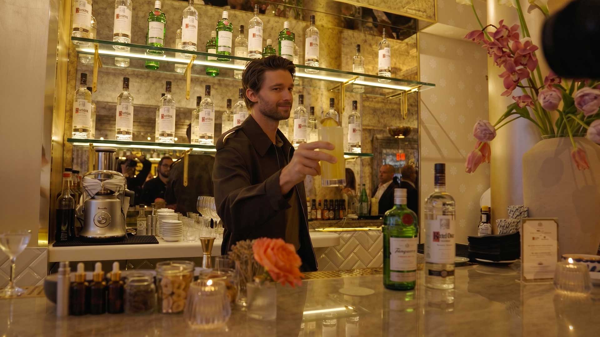 Patrick Schwarzenegger holding up a yellow cocktail glass behind a bar with bottles and flowers on the counter.