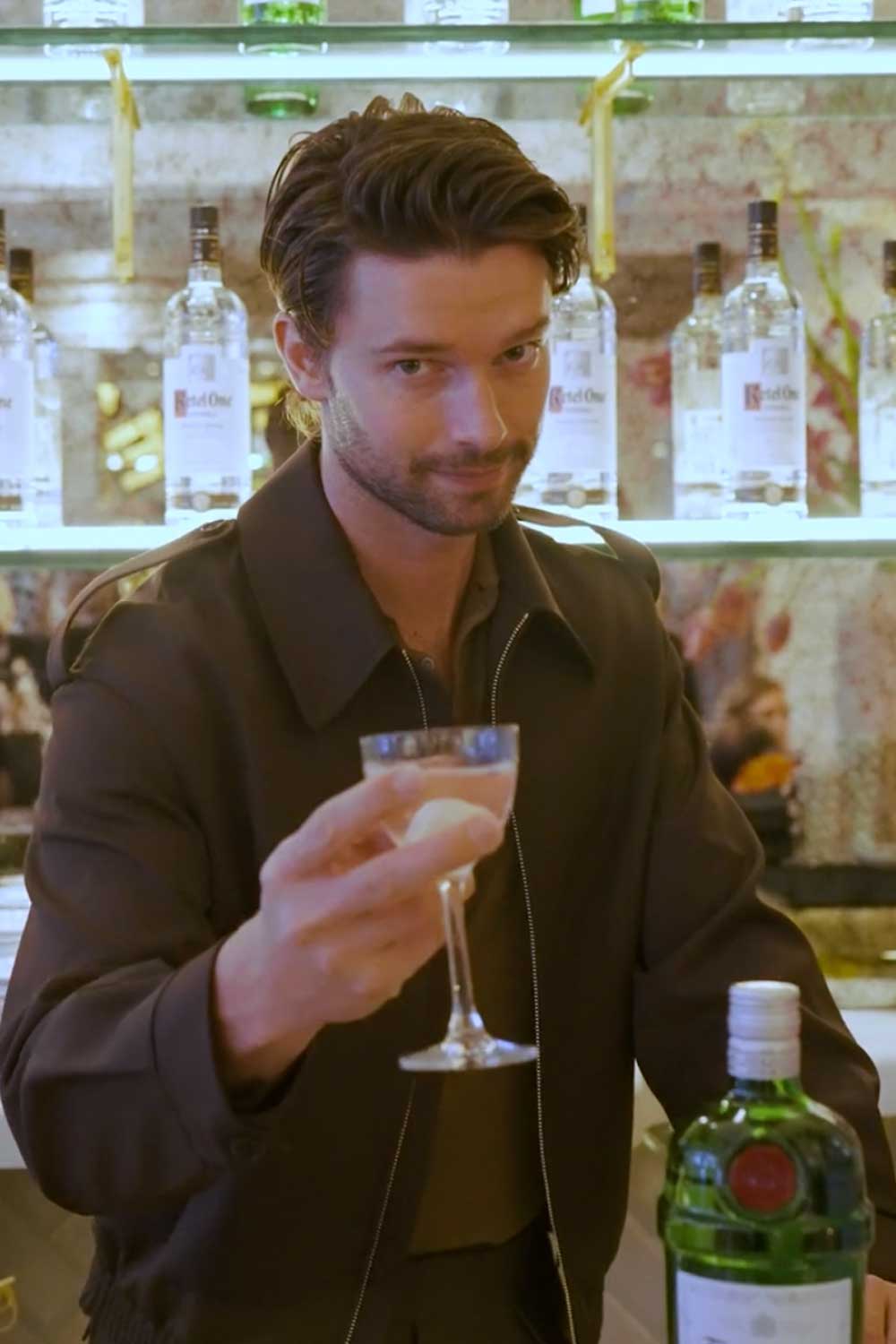 Patrick Schwarzenegger with brown hair and a jacket, holding a cocktail glass, standing in front of liquor bottles on shelves.
