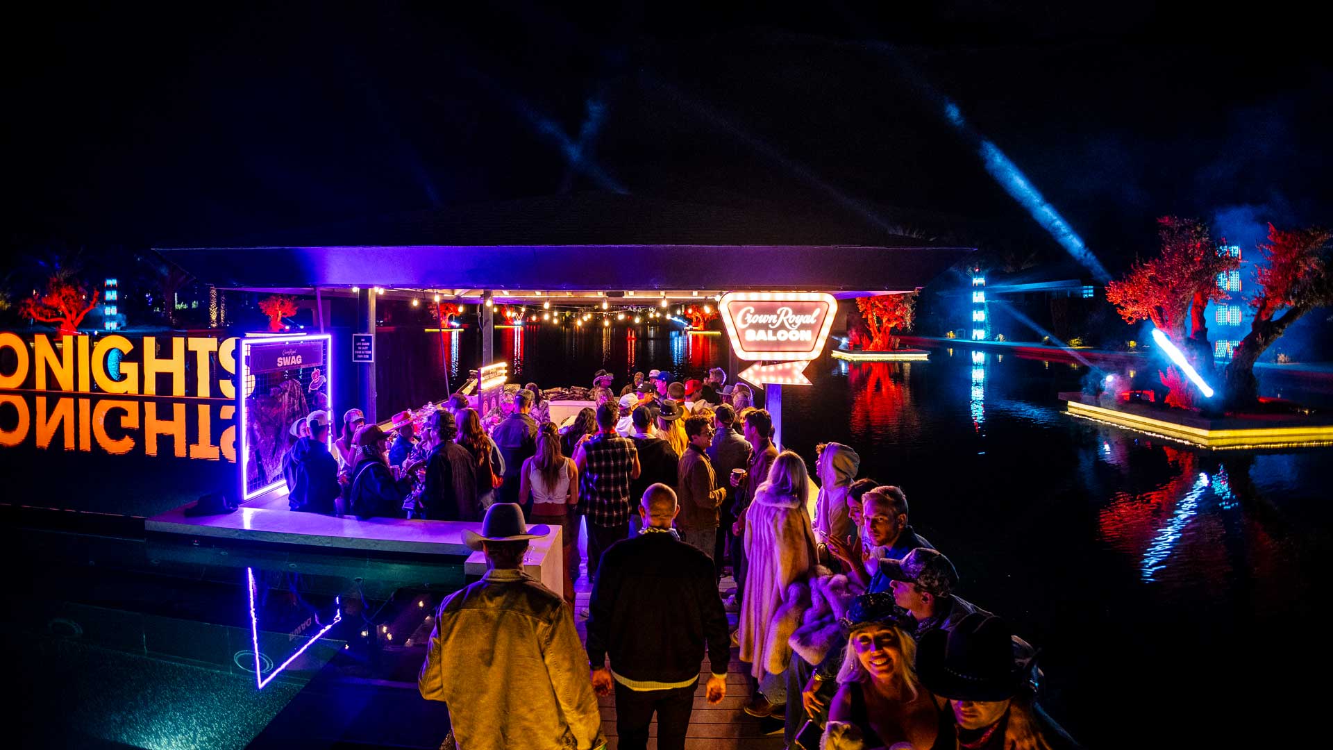 Crowd of people gathered at a brightly lit outdoor nighttime event by water with neon lights and a Crown Royal Saloon sign.