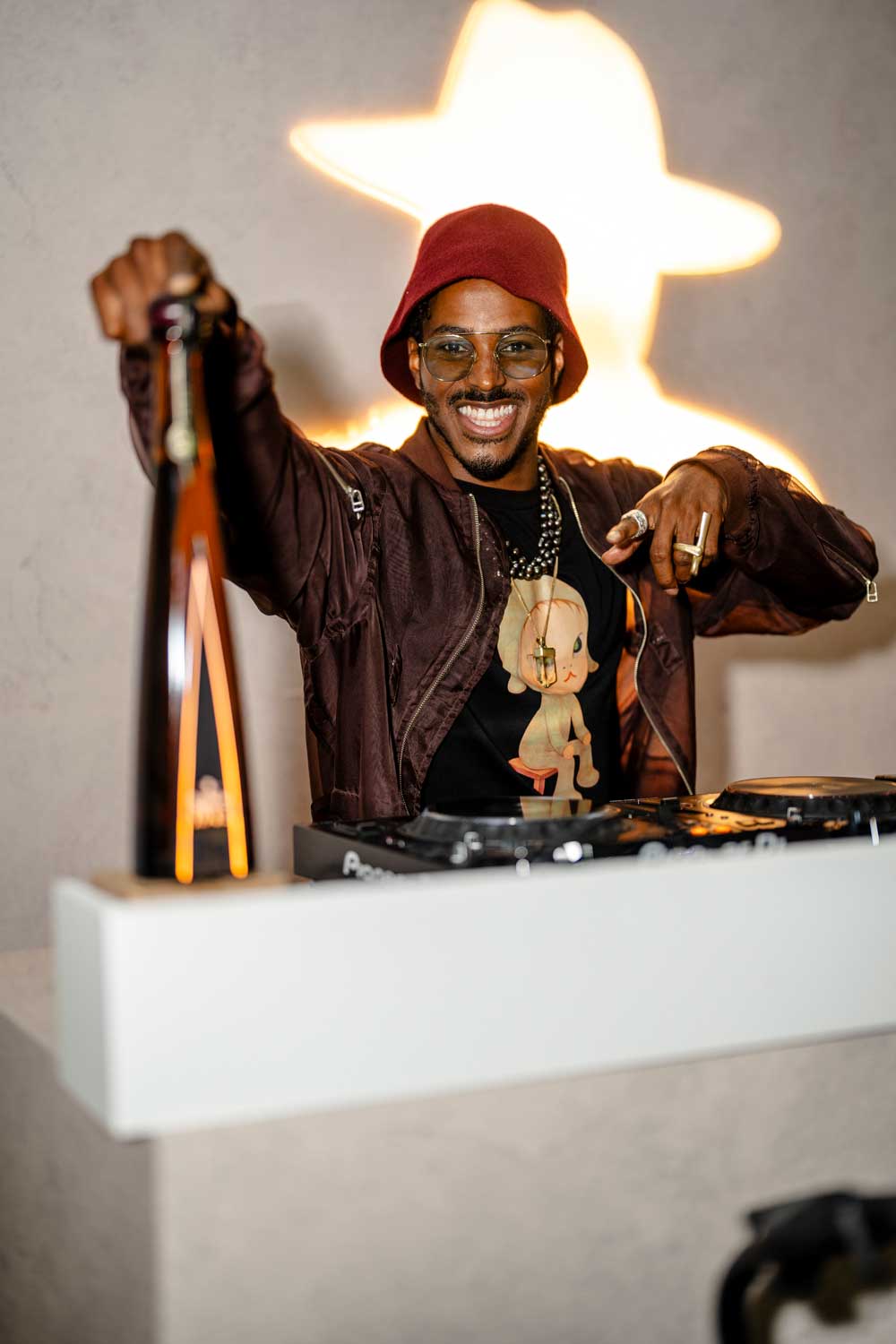 Smiling DJ in a red hat and glasses holds up a bottle behind a DJ deck with a glowing star-shaped light.