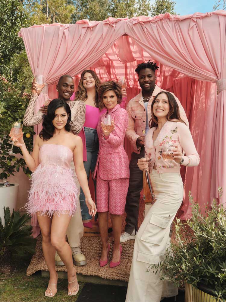 Group of six diverse people dressed in pink and beige holding drinks in front of pink draped backdrop.