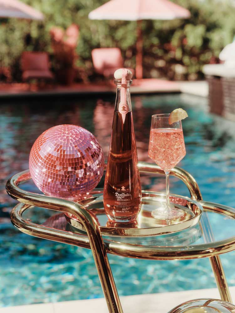 Bottle of Don Julio Rosado tequila, a glass with a cocktail and lime, and a pink disco ball by the pool.