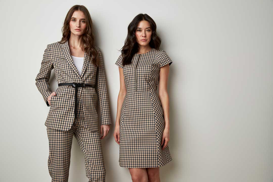 Two women standing against a plain light gray background wearing matching beige and black checkered outfits; the woman on the left wears a belted blazer and trousers with a white top underneath, the woman on the right wears a fitted short-sleeve dress with a vertical black zipper detail on the front.
