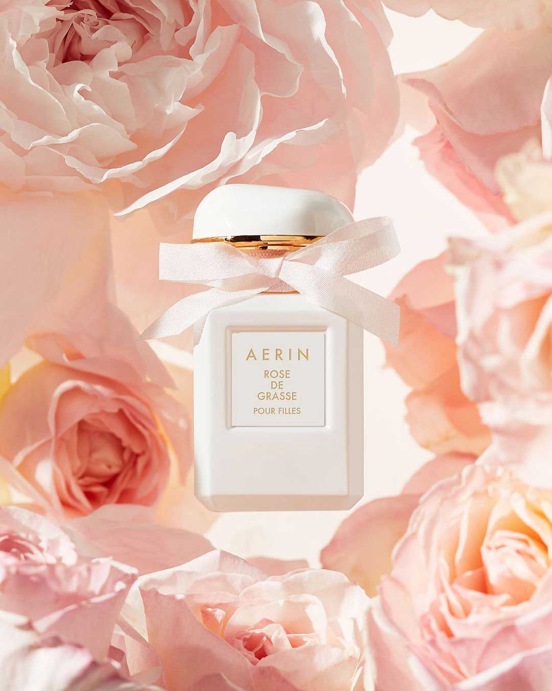 Aerin Rose de Grasse Pour Filles perfume bottle with a white bow around its neck, surrounded by soft pink and peach roses.