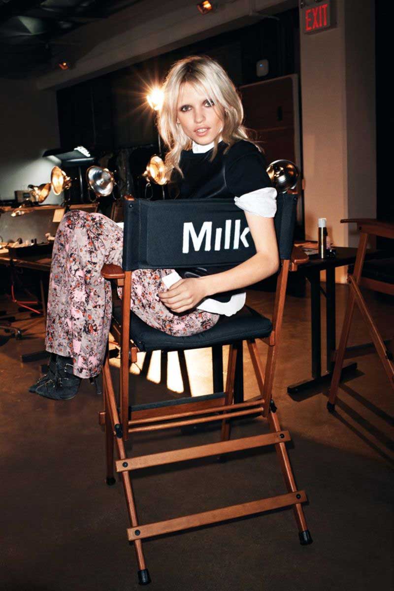 Blonde woman with layered hair and smoky eye makeup sitting sideways on a director's chair labeled 'Milk' in a dimly lit room with studio lights and an exit sign in the background.