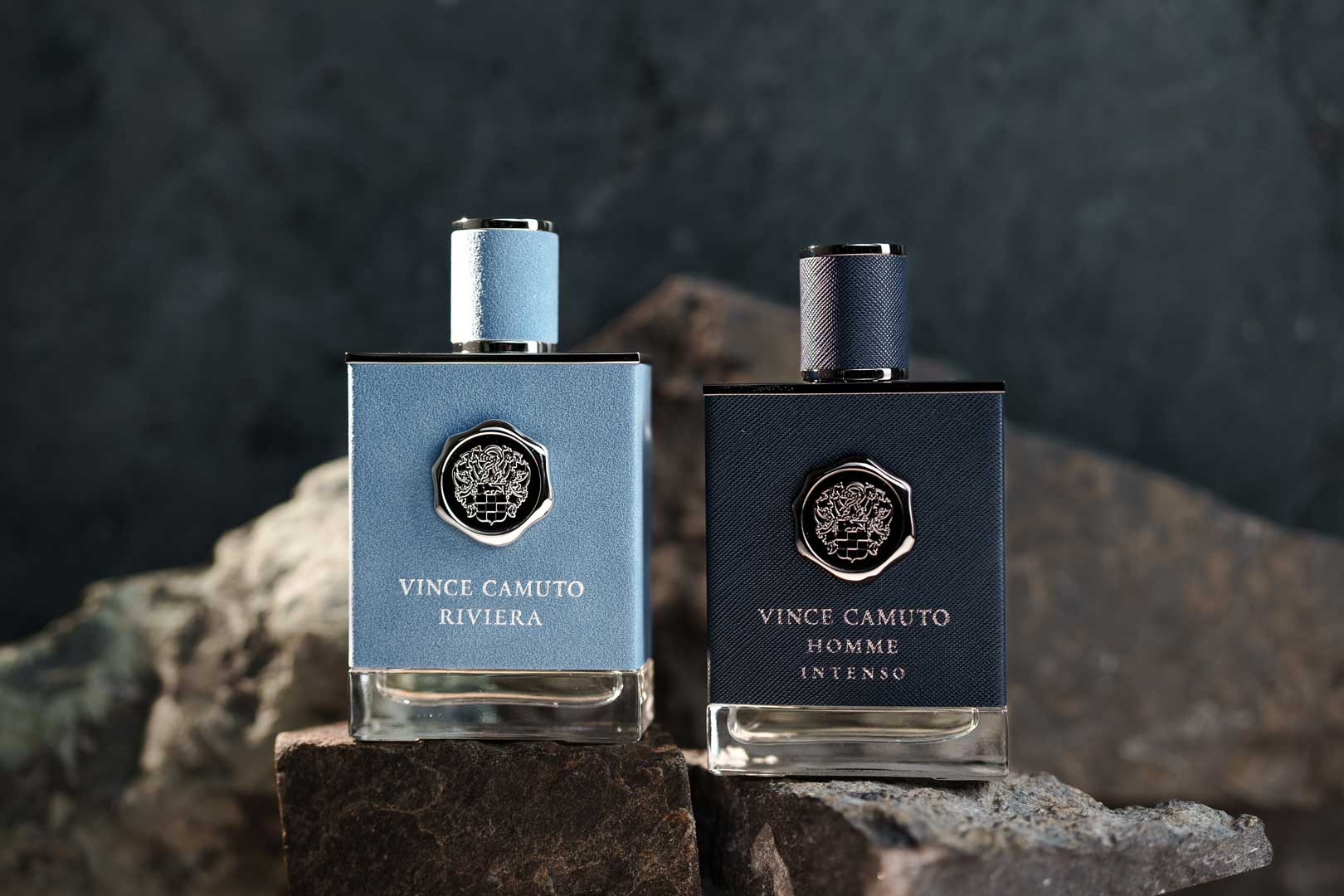 Two Vince Camuto cologne bottles placed on rocks; the left bottle is light blue labeled 'Riviera,' and the right bottle is dark blue labeled 'Homme Intenso,' both with a silver emblem.