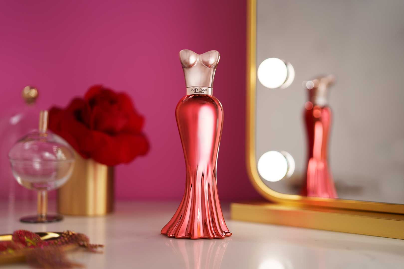 Red metallic perfume bottle shaped like a woman's torso in a form-fitting dress, labeled 'Ruby Rush' by Paris Hilton, placed on a white surface with a mirror and lights behind it, and blurred red flowers and a glass container in the background.