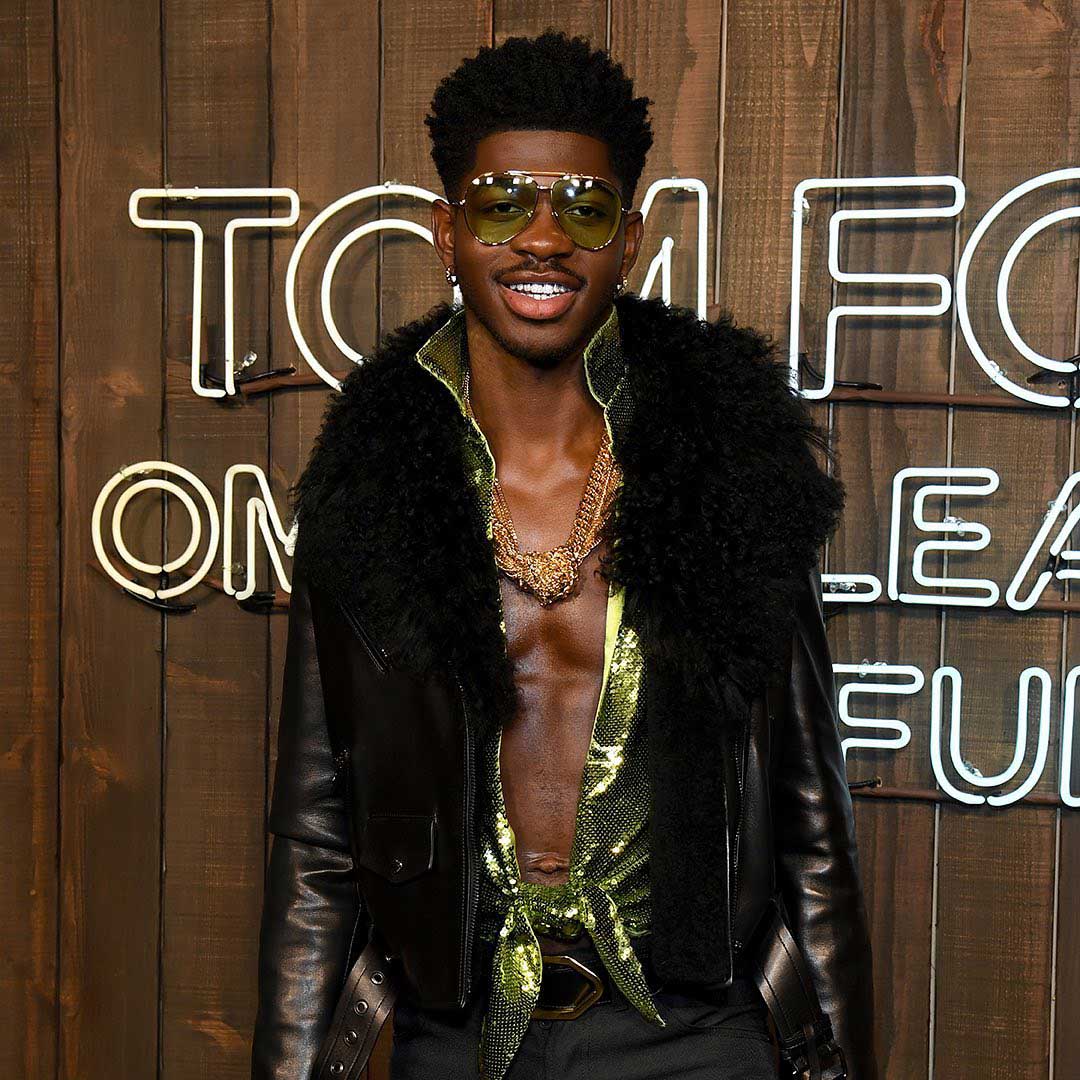 Lil Nas wearing yellow-tinted aviator sunglasses, a black leather jacket with a large black fur collar, and a shiny green shirt tied at the waist with gold chains around his neck, standing in front of a wooden wall with illuminated white neon text.