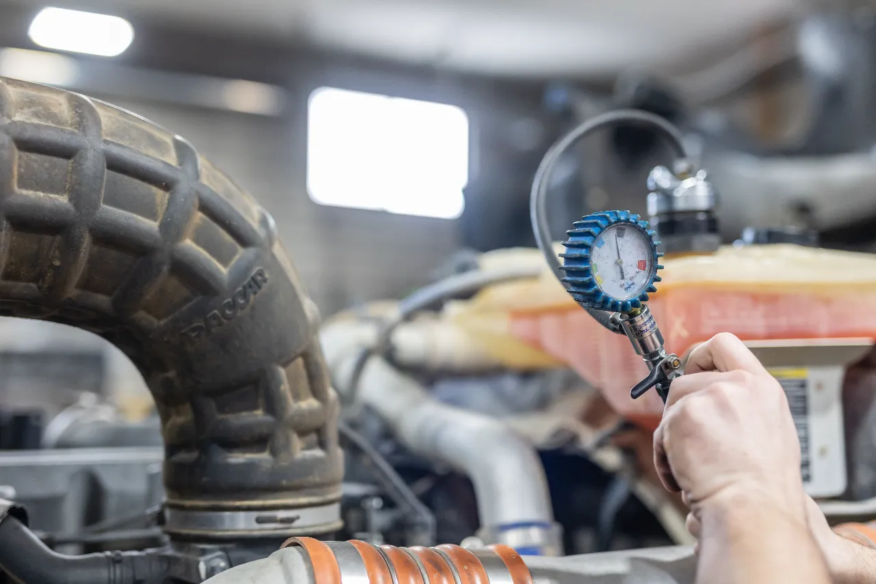 Mechanic checking engine pressure with gauge near PACCAR intake hose and coolant reservoir.