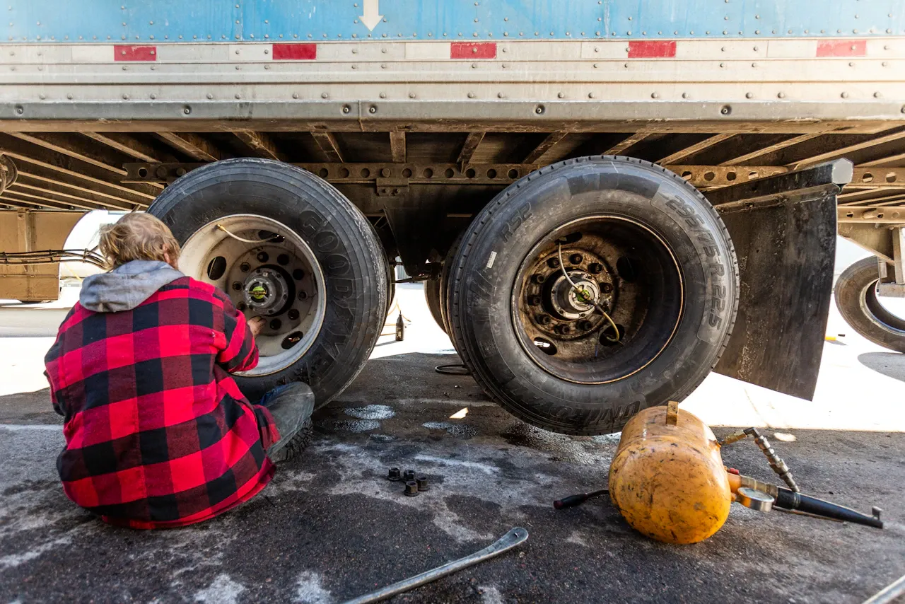 Roadside technician services a semi-trailer wheel hub with tools and an air tank beside dual Goodyear tires on pavement.