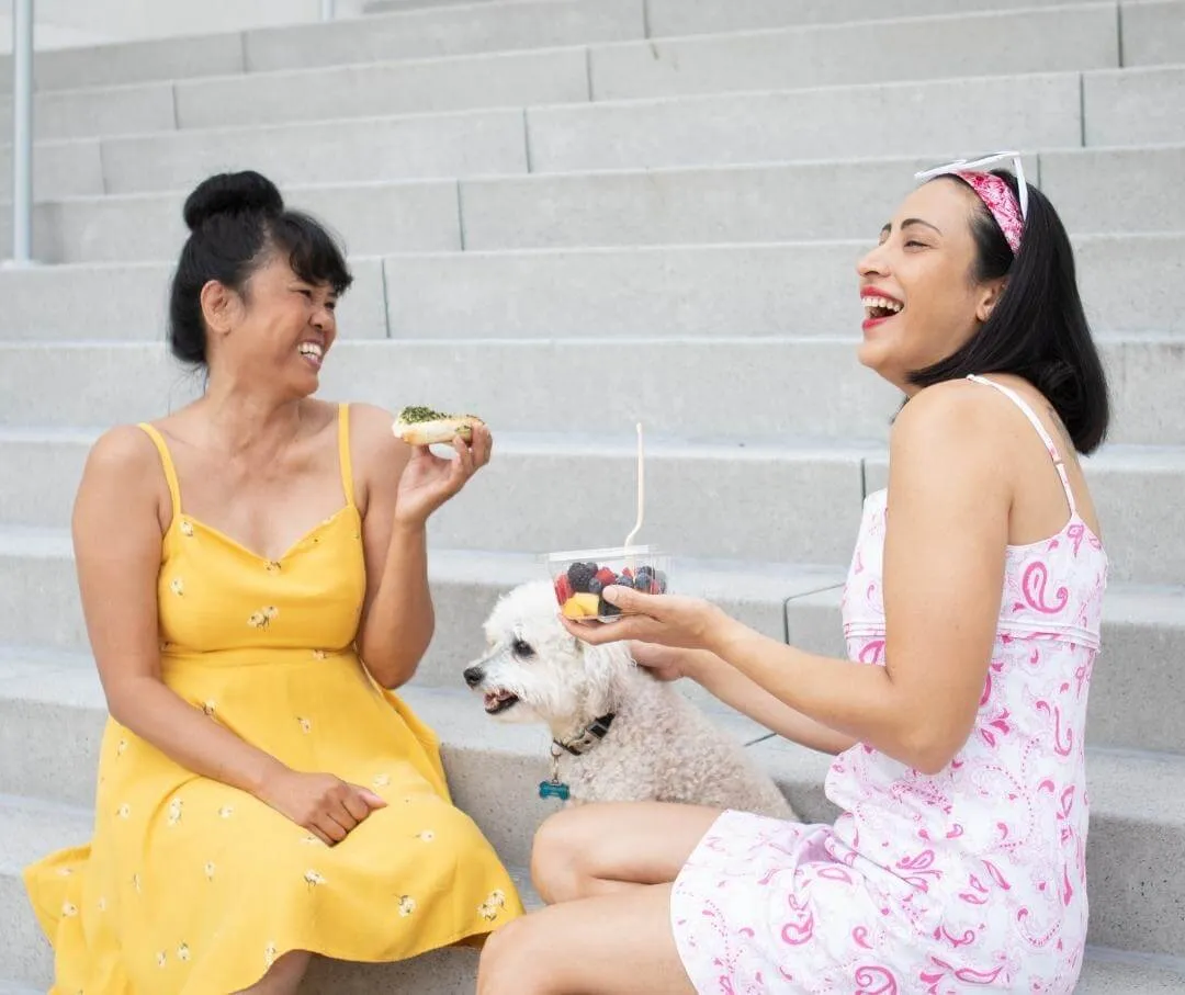 women sitting on some steps outdoors with a small white dog, eating food and laughing