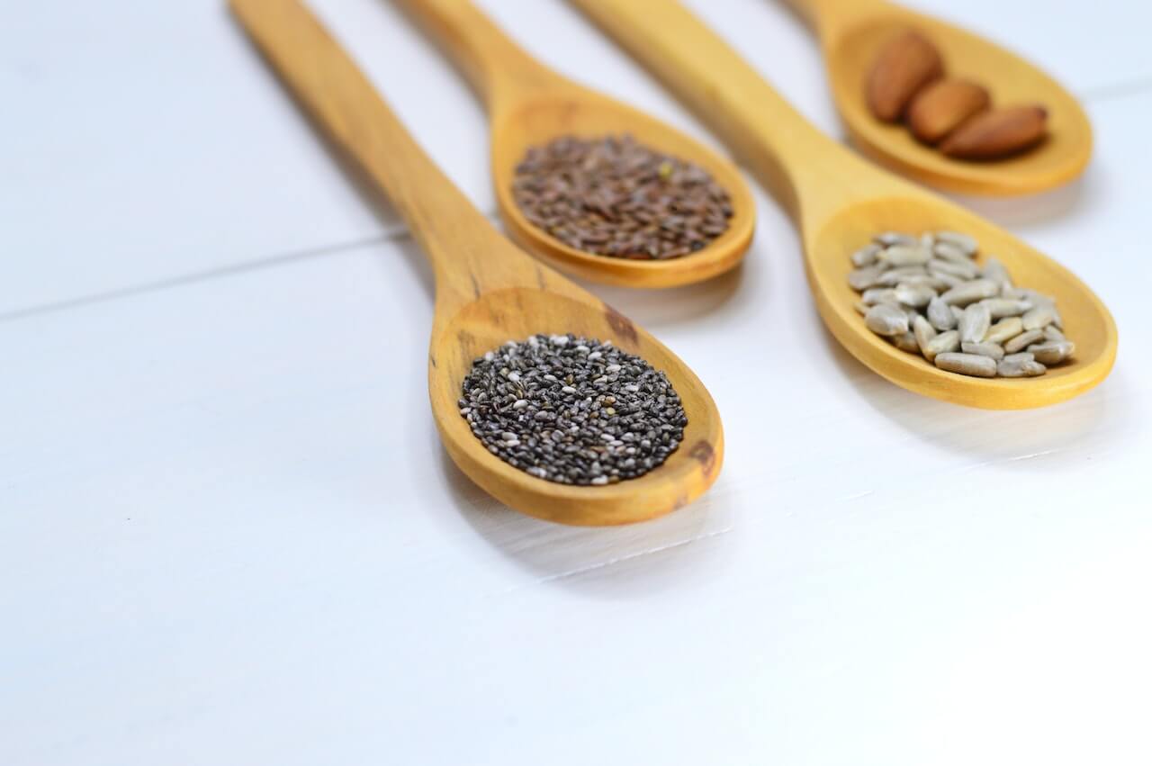 Four wooden spoons with different seeds: chia, flax, sunflower, and almonds