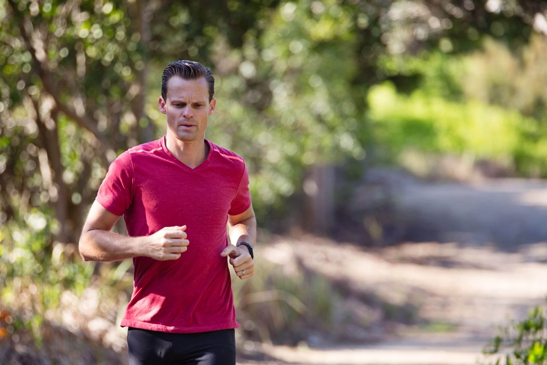Runner in red shirt jogging on a tree-lined trail with focused expression