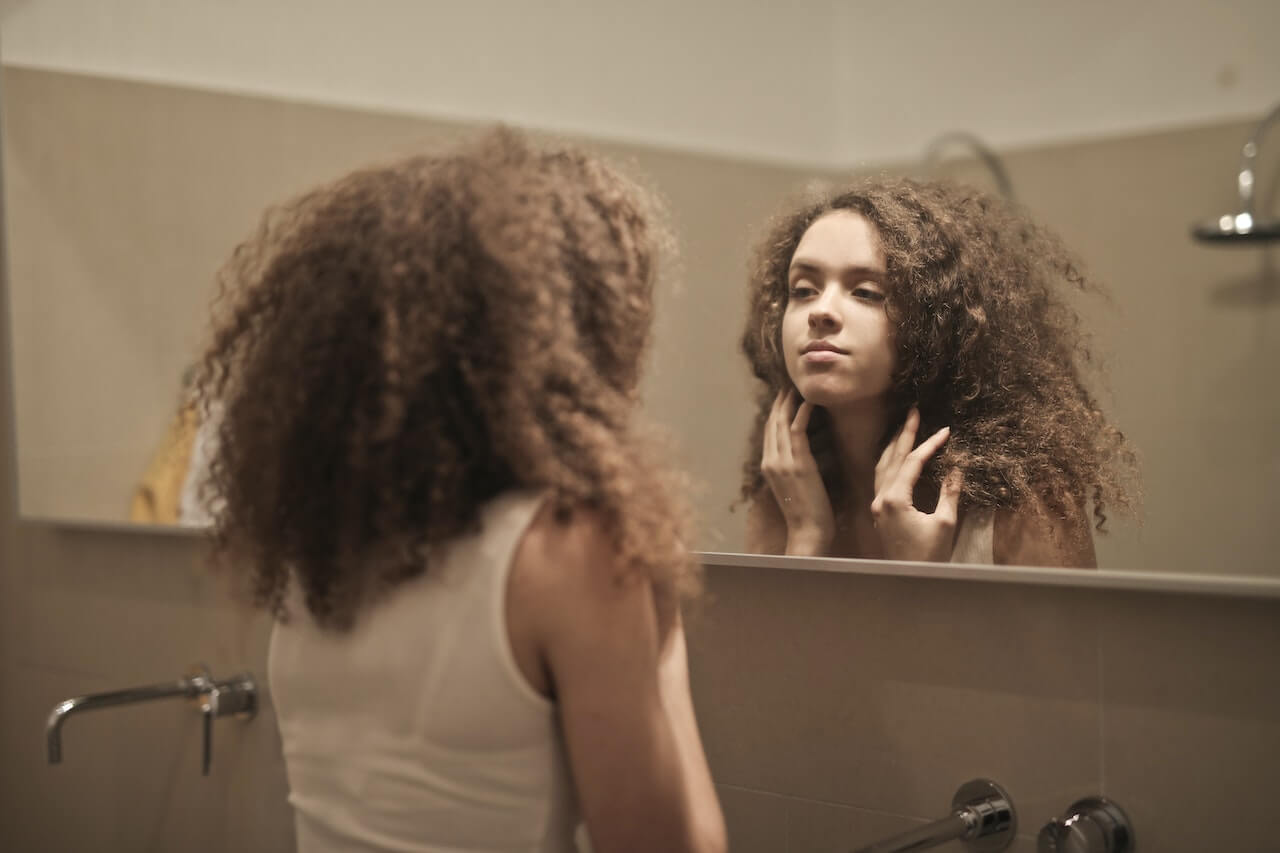 Person with curly hair looking thoughtfully at reflection in bathroom mirror