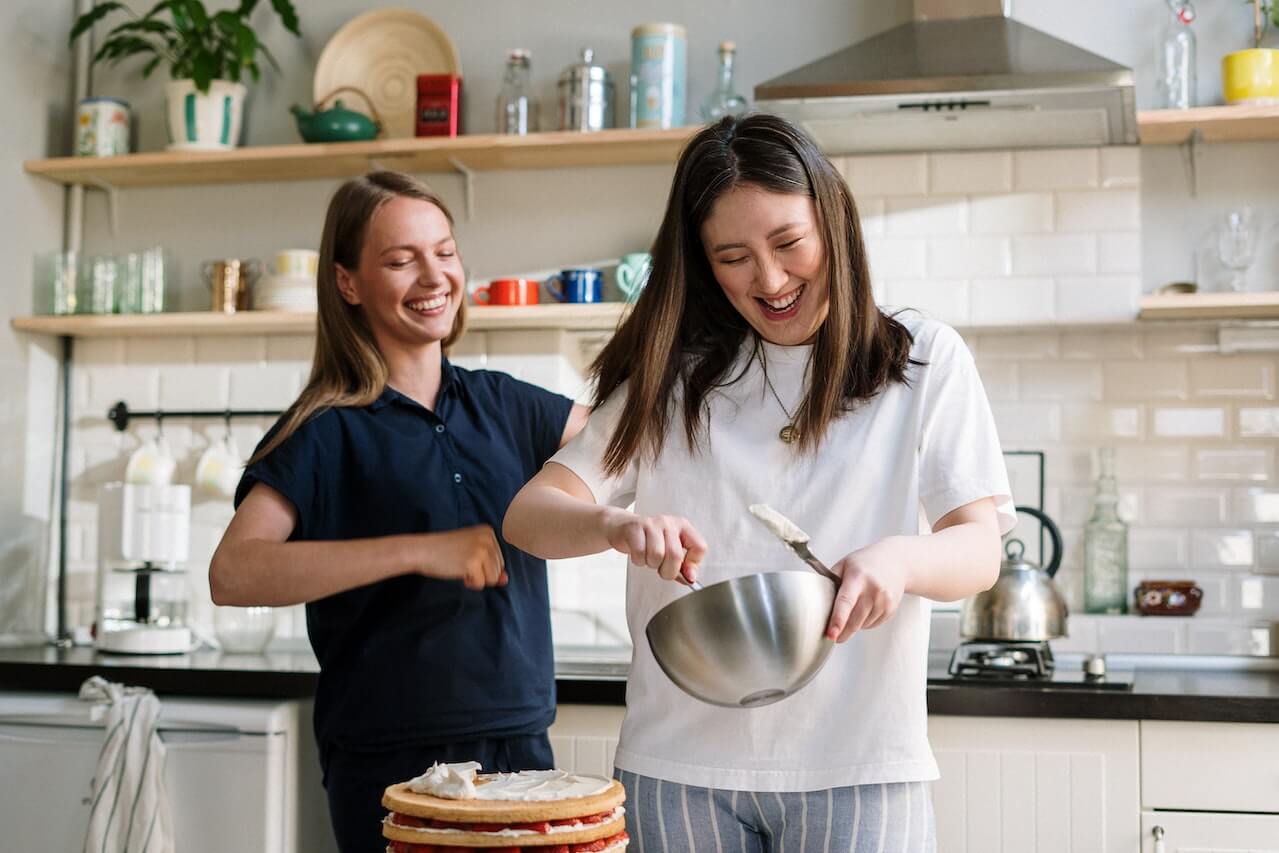 Two friends laughing while baking a layered cake in a kitchen