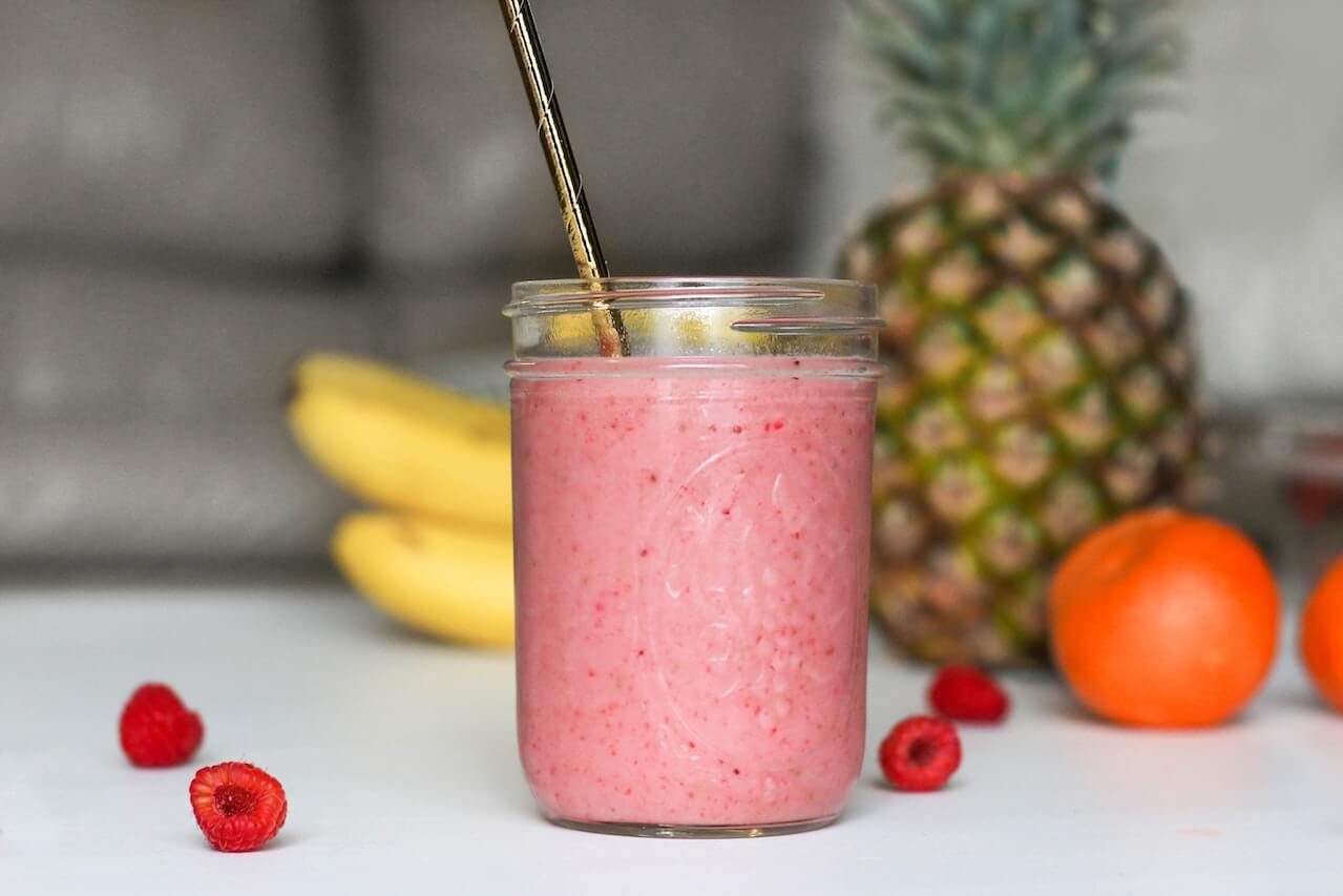 Pink smoothie with raspberries, banana, pineapple, and orange in background