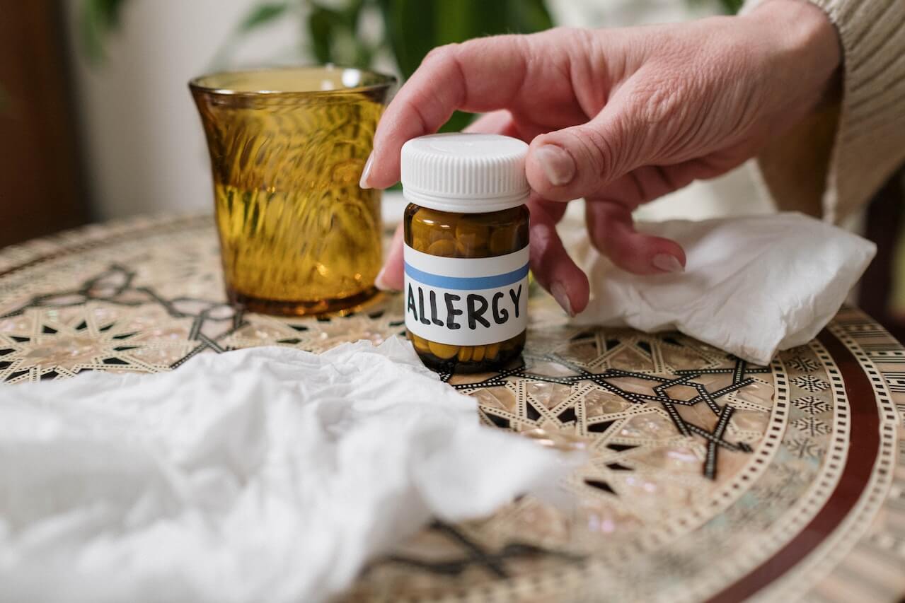 Hand holding allergy medication bottle with tissues and yellow glass
