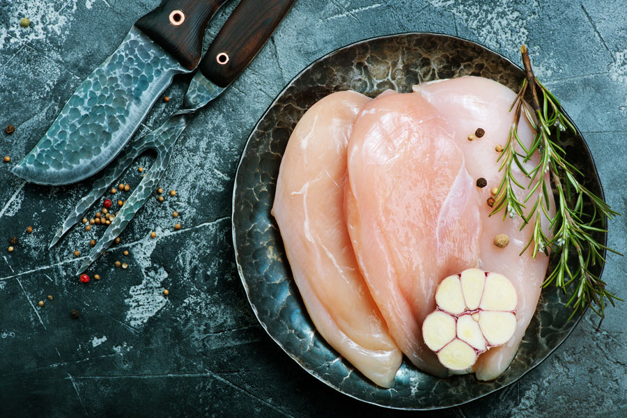 Raw chicken breasts on plate with rosemary, garlic, and knife on blue surface.