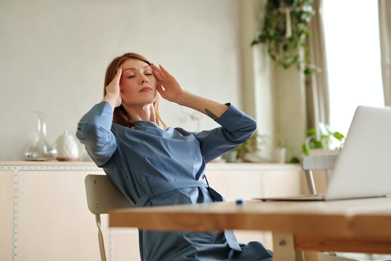 Woman in blue shirt experiencing stress or headache at office desk with laptop