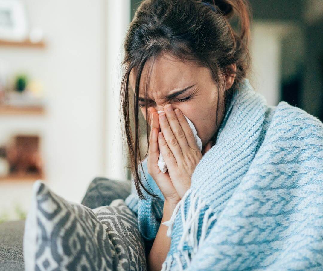 Woman sneezing into tissue while wrapped in blue blanket at home.