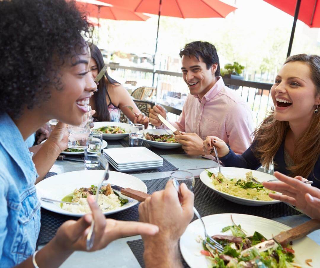 Group of friends laughing and enjoying salad meal together under red umbrellas outdoors