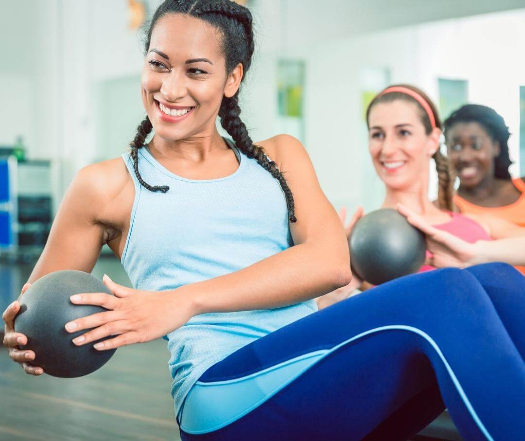 Woman holding medicine ball doing sit-ups in gym class with instructors