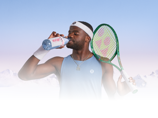 Frances Tiafoe in blue Lululemon tank top and white headband holds a green tennis racket and a bottle of Evian water against snowy mountain background.