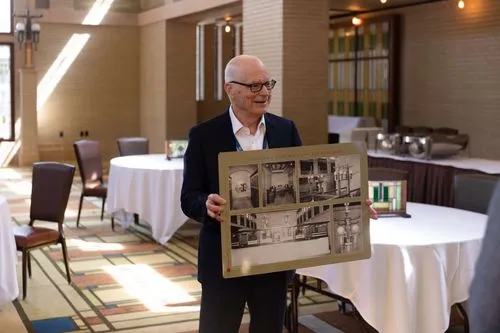 In the image, The Honorable Bill Schickel, who is the Mayor of Mason City, is holding a poster with historical photographs of the Historic Park Inn and City National Bank. His expression is pleasant and he seems to be engaging with an audience, during a tour within the ballroom.