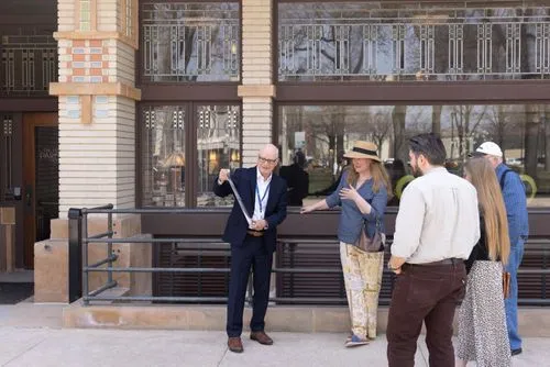 
The image features a group of people on a walking tour outside the Historic Park Inn Hotel. The group is led by a man identified as Mason City Mayor, gesturing informatively to his audience, which includes an artist and author among the attendees. They appear engaged, standing in front of the hotel’s distinct architectural facade with its decorative windows.