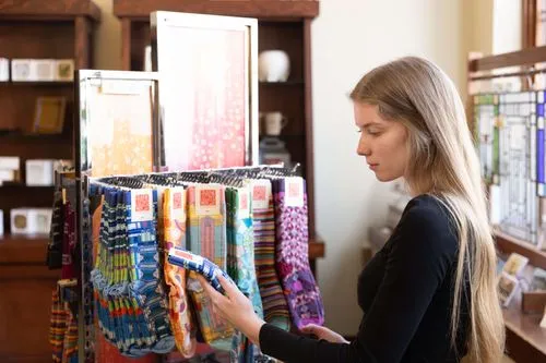 The photo shows a woman in the Historic Park Inn gift shop examining a variety of colorful socks displayed on a rotating rack. The designs on the socks are vibrant and likely inspired by architectural or artistic motifs. The shop has a classic feel with wooden shelves and a selection of decorative items and souvenirs in the background, catering to those appreciating design and artistry.