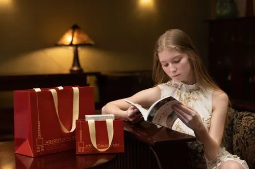 The photo shows a young woman seated in the Skylight room, deeply engrossed in reading a book that is available for purchase in the gift shop. Next to her are the distinctive Inkfish-designed shopping bags, featuring the logo of "Wright on the Park." The setting exudes a calm atmosphere, with soft lighting from a table lamp contributing to the serene environment suitable for reading and relaxation.