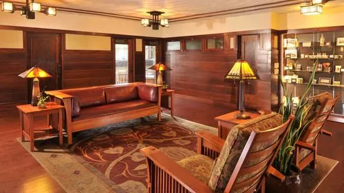 
This image captures the Mahogany Room next to the original bank offices, featuring dark wood panels and trim that evoke a warm, historical ambiance. Leather furniture, including a long sofa and patterned upholstered chairs, offers comfortable seating. Accent tables with stained glass lamps provide soft lighting, complementing the natural light from the windows. Displays of small items and books add a personal touch to the room, which is finished with an ornate floral carpet, enhancing the stately atmosphere of this well-preserved architectural space.