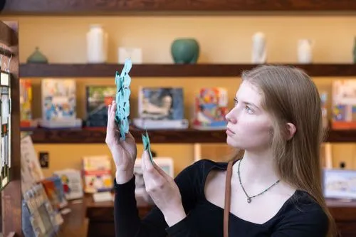 The image depicts a young woman in a gift shop, closely examining a delicate, vertically-hanging mobile with aqua-colored abstract pieces. The mobile appears to be a decorative art piece, and the woman is holding it up to get a better look, possibly appreciating its design and how it balances. She is surrounded by shelves stocked with a variety of artistic and architectural-themed items, suggesting a focus on design and aesthetics in the merchandise selection.