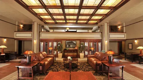 The photo depicts the skylight room of the Historic Park Inn Hotel, showcasing a cozy lobby area. The room is illuminated by a large, grid-patterned skylight that casts a warm glow onto the space below. Comfortable leather sofas and chairs are arranged around a central coffee table, creating a welcoming sitting area. Wood paneling and trim add to the classic charm of the room, while elegant table lamps and wall-mounted light fixtures provide additional warmth. The décor is consistent with Frank Lloyd Wright’s design aesthetic, featuring geometric lines and natural colors.