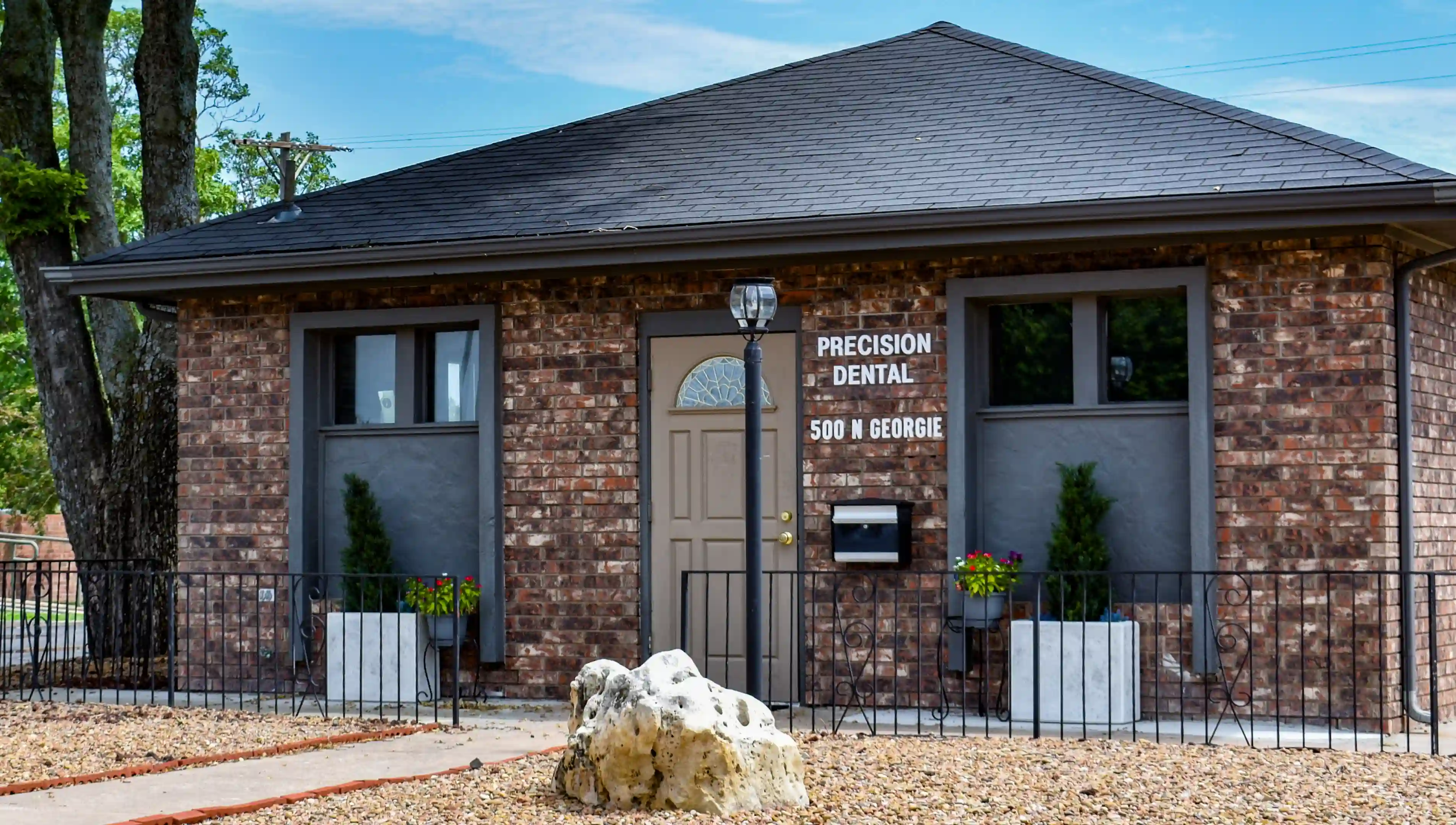 Brick building with a gray door and two windows, sign reading Precision Dental 500 N Georgie, small plants in planters, and a lamp post in front.