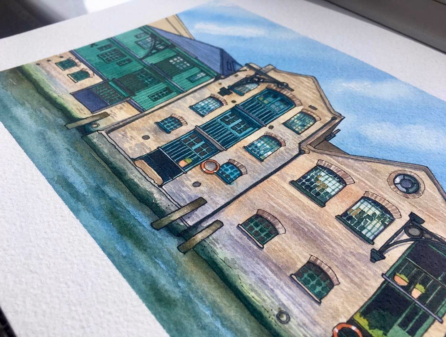 An angled close up view of a painting depicting a row of old English industrial buildings on a waterfront in London. The buildings have warm brown brickwork, dark green wooden cladding, plenty of windows and balconies. The orange life rings stand out against the brickwork.