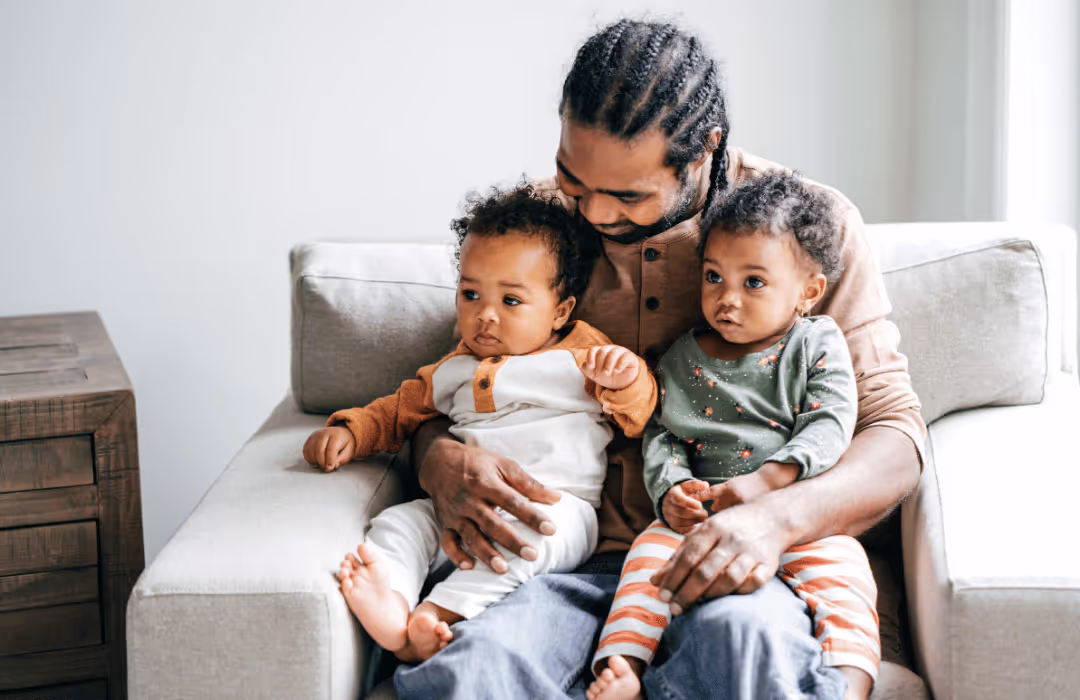 Man with braided hair sitting on a beige sofa holding two toddlers, one in a brown and white outfit and the other in a green top with striped pants.