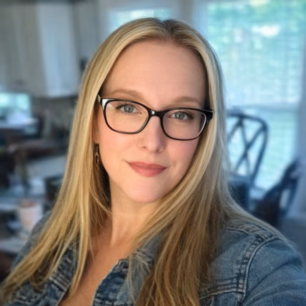 Close-up of a smiling woman with long blonde hair and glasses wearing a denim jacket indoors.
