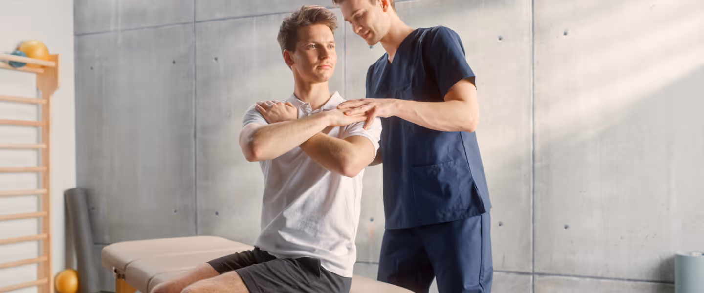 Physical therapist assisting a seated man with arm cross stretch in a therapy room.