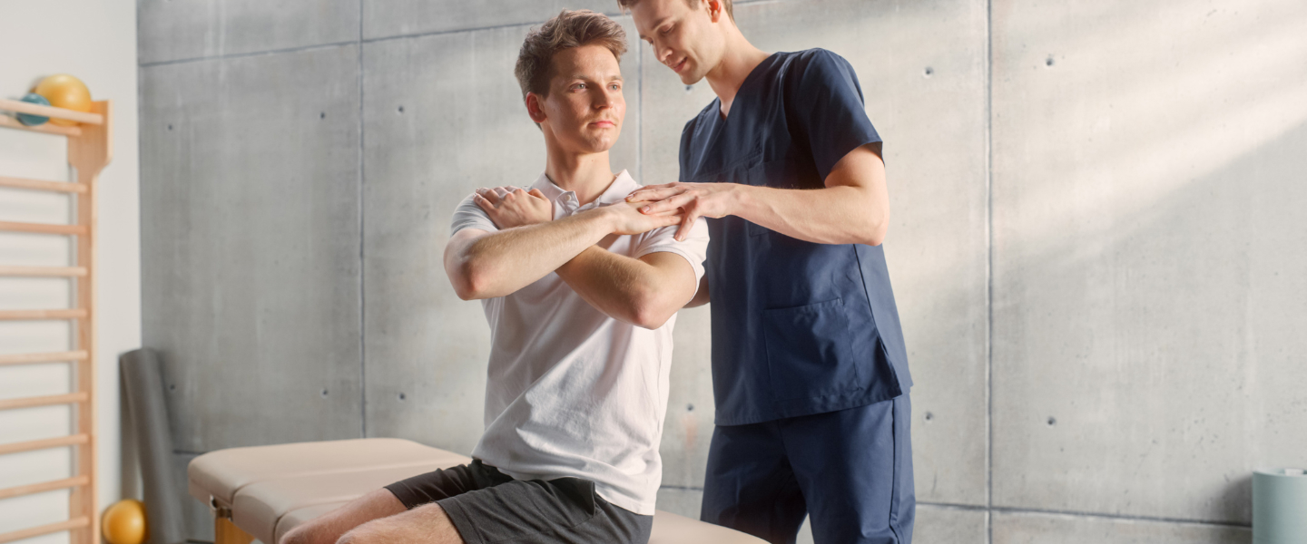 Physical therapist assisting a seated man with arm cross stretch in a therapy room.