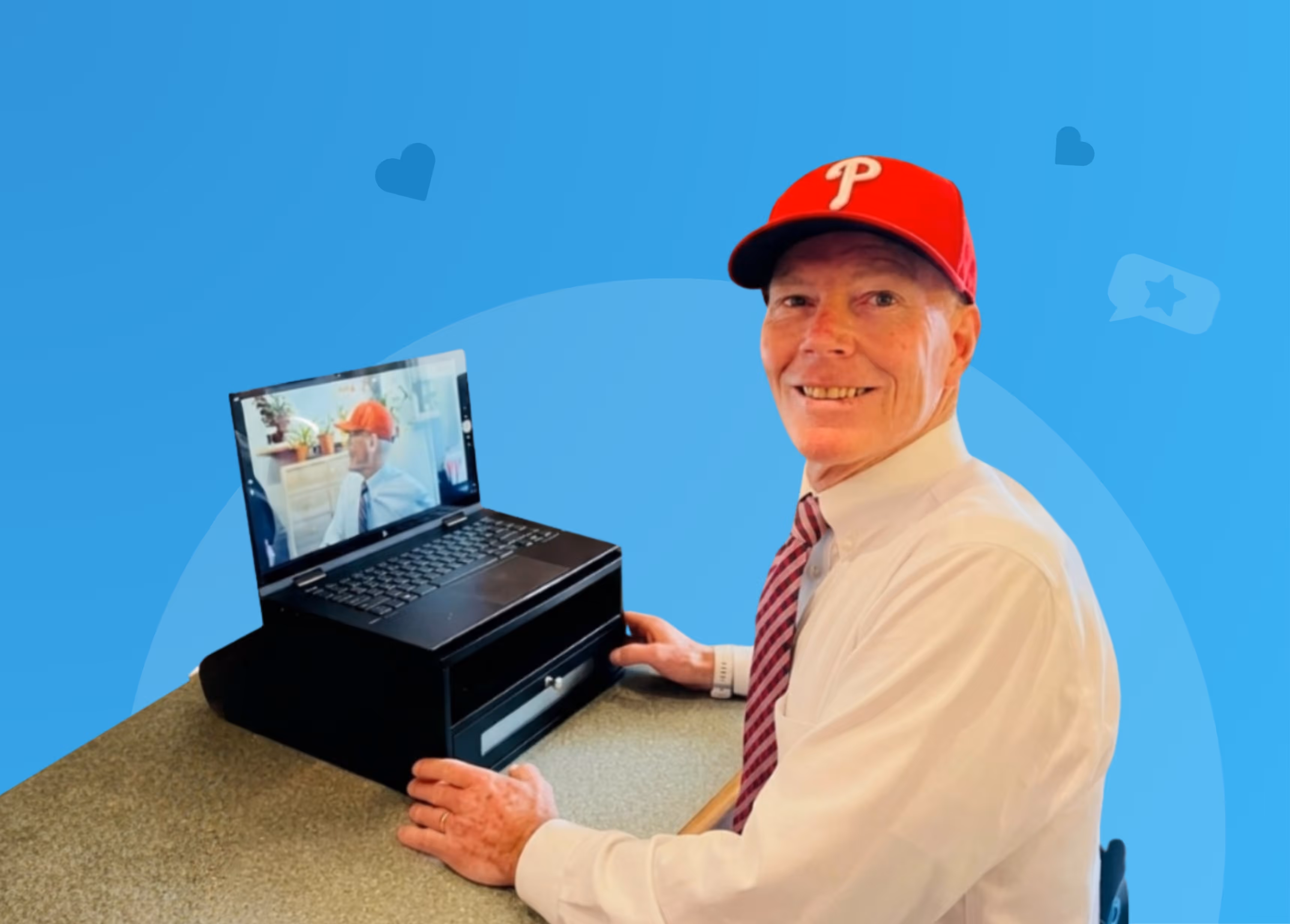 Smiling man wearing a red Philadelphia Phillies cap, white shirt, and striped tie, sitting at a table with a laptop showing his own image.