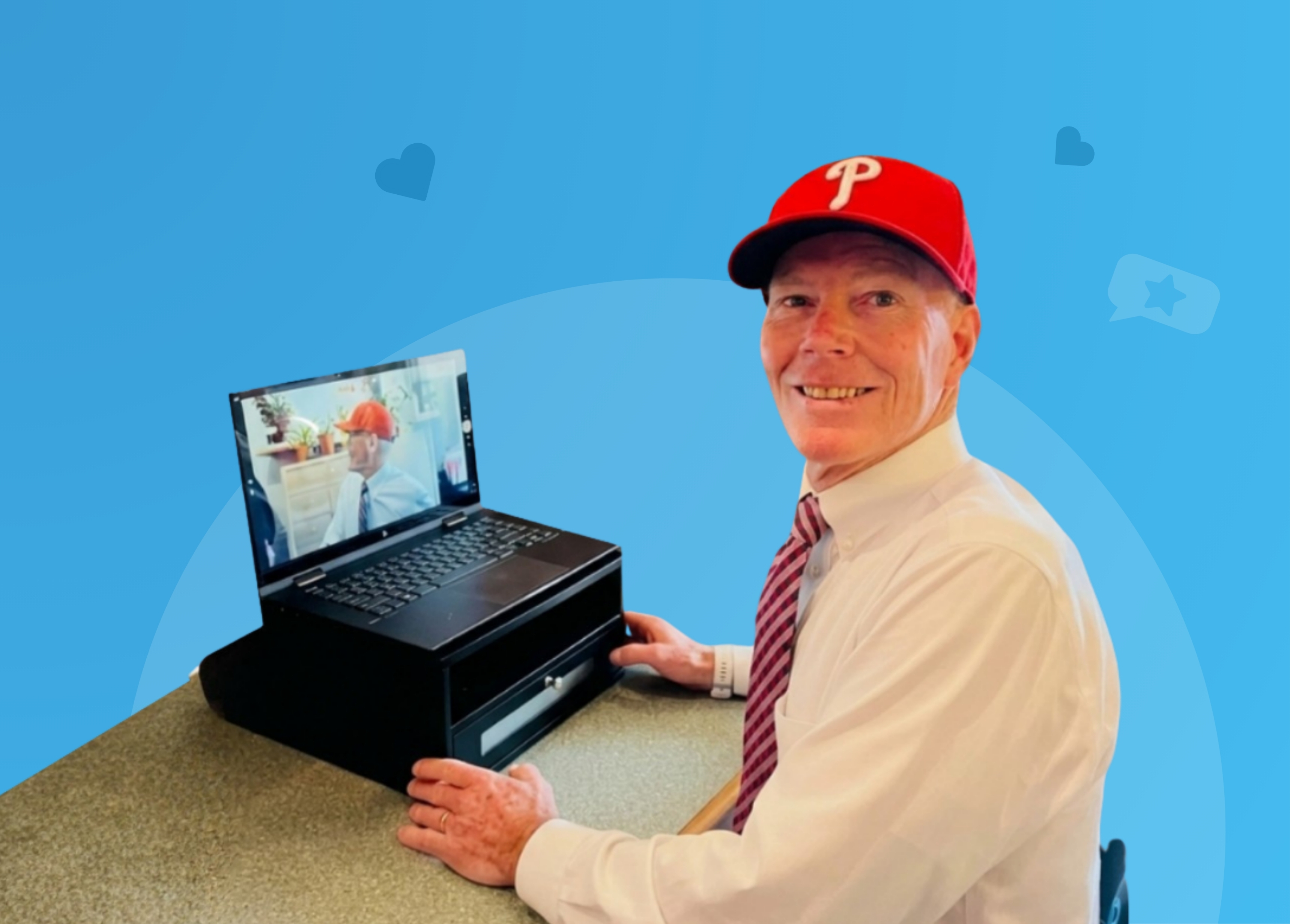 Smiling man wearing a red Philadelphia Phillies cap, white shirt, and striped tie, sitting at a table with a laptop showing his own image.