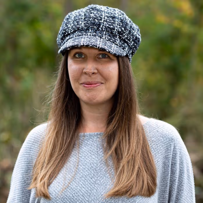 Smiling woman with long brown hair wearing a gray knit sweater and a black-and-white cap outdoors.