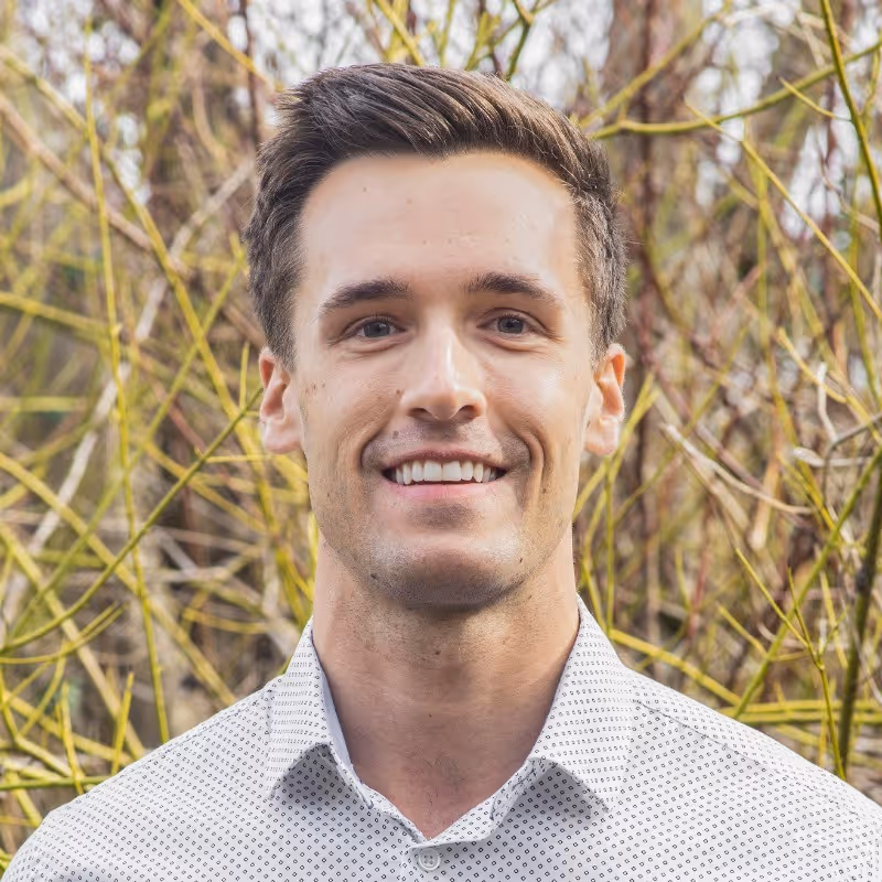 Smiling man with short brown hair wearing a light patterned shirt, standing outdoors with leafless branches behind.