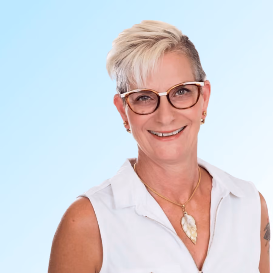 Smiling woman with short blond hair wearing glasses and a white sleeveless top with a leaf necklace.