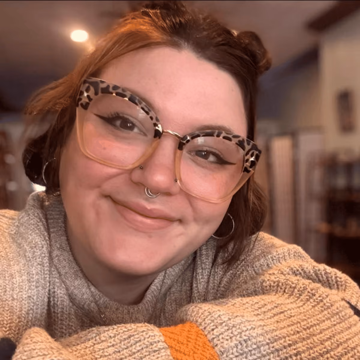 Smiling person with glasses, septum piercing, and cozy knit sweater in a warmly lit indoor setting.
