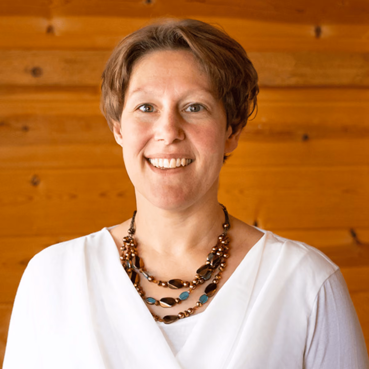 Smiling woman with short brown hair wearing a white top and a layered necklace, wooden background.