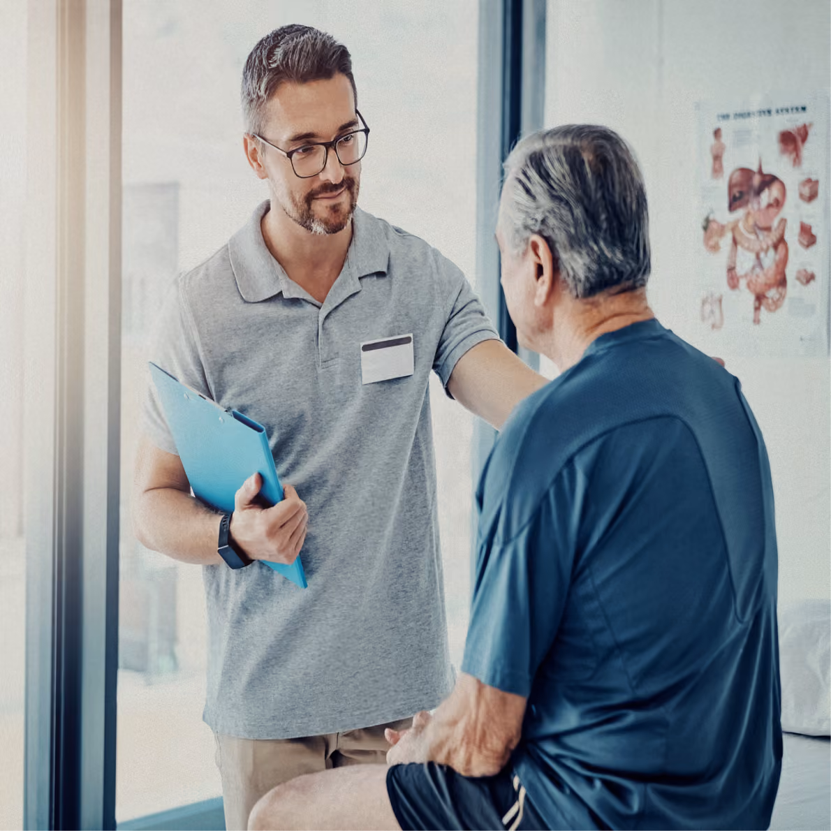 Male healthcare professional with glasses holding a blue clipboard talks to an elderly man seated in a medical clinic with anatomy poster on the wall.